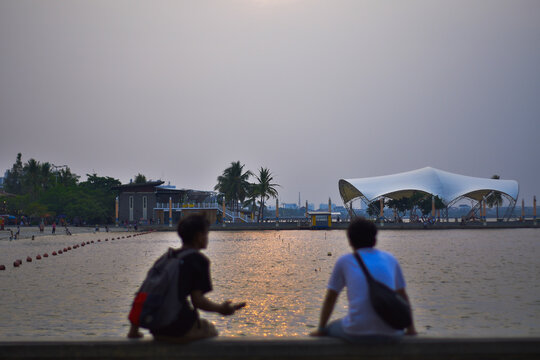 Two people sitting back to back while looking at the beach and sunset in Ancol, Jakarta, October 14, 2024