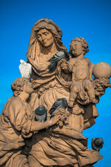Obraz premium Statue with mother and child on Charles Bridge in Prague surrounded by pigeons on a clear day