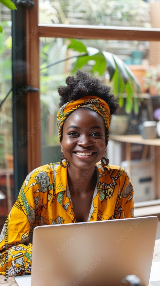 Wall mural Vibrant young woman in yellow floral attire and headwrap smiles brightly while working on laptop in sunny, plant-filled space, embodying joy and productivity.