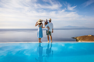 A family on summer holiday time stands at the edge of an infinity swimming pool and enjoys the view over the ocean