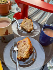 A patio table with a mug of tea, a cup of coffee, and two plates of coffee cake. The sweet bread is on clear glass plates. A red patio chair with a green striped cushion is next to the food.