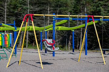 A children's playground with a colorful metal swing set, a children's swing chair and a monkey climber. The background is a wooden fence and a wooded area. A climbing structure made of green metal. 