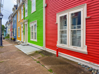 Street view of multiple colorful wooden buildings of various bright colors; red, green, yellow, and blue. The small structures have double-hung windows and white doors. The sidewalk is concrete.