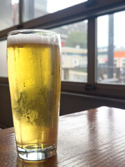 A tall clear pint beer glass filled with cold foam from a lager ale. The golden colored ale sits on the edge of a wooden patio table at a microbrewery. There's foam on top of the chilled glassware.