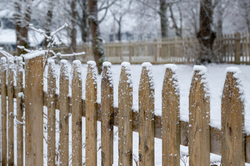 A natural wood picket fence with the tips of the palings covered in fresh white snow. The divider has a snow covered grassy garden with large trees in the yard. The fence encloses the property.