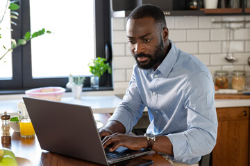 A focused African-American man works on his laptop at a kitchen counter. The modern kitchen is well-lit, with fresh fruit and a glass of juice nearby, creating a productive atmosphere.