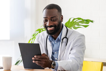 A smiling African-American male doctor in a white coat, with a stethoscope around his neck, uses a tablet in a bright and modern office. He appears engaged and happy, providing medical care or consult