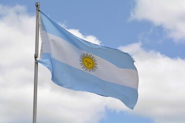 The national flag of the Argentine Republic flies up a flagpole on a beautiful summer day with a background of blue sky and white clouds.