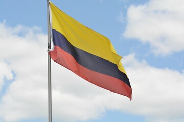 The national flag of Colombia flies up a flagpole on a beautiful summer day with a background of blue sky and white clouds.