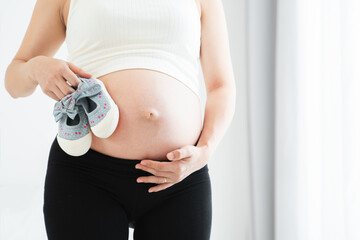 Pregnant Asian young woman standing and holding a little baby shoes near her belly with love and care at home near window. Pregnancy lifestyle. Copy space.