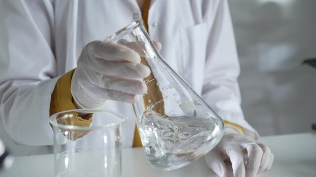 Unknown scientist wearing white protective gloves is carefully shaking up a clear chemical solution inside an Erlenmeyer flask in a laboratory, close up. Science and medicine