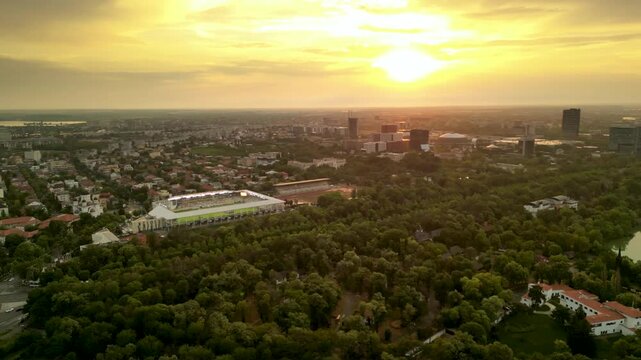 High angle drone footage of Herastrau and Arcul de Triumf in Bucharest Romania. Green park shot on a sunny day during golden hour. Cityscape of warm summer evening. Rugby stadium visible from above
