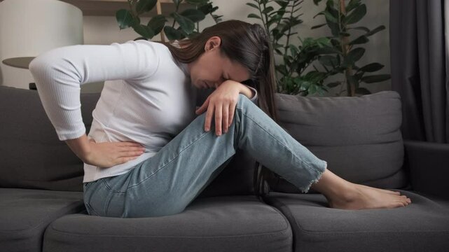 Flatulence young woman hand in stomach ache, suffer from food poisoning, abdominal pain and colon problem, gastritis or diarrhoea sitting on couch at home. Patient belly, abdomen or inflammation