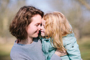 Fototapeta premium Young beautiful mother and an adorable blond little daughter are hugging and kiss each other in a park, happy family
