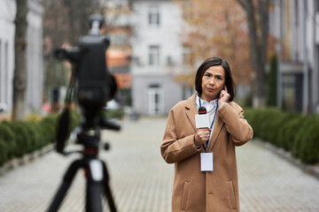 A multiracial female journalist in a beige coat prepares to deliver a news report in an autumn urban landscape.