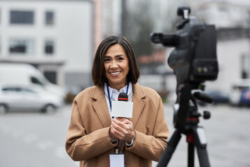 A multiracial female journalist in a beige coat reports live outdoors, sharing vital news through...
