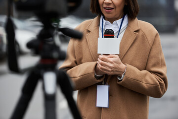 A multiracial female journalist in a beige coat holds a microphone while preparing to report from a...