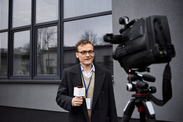 A professional journalist in glasses speaks into a microphone while standing near a camera outdoors.