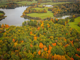 An aerial view of a forest transitioning into autumn, with green, orange, and yellow trees surrounding a calm lake. The landscape includes grassy fields, winding water bodies