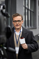 A professional journalist in a coat speaks into a microphone, delivering news updates outdoors.