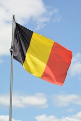 The national flag of Belgium flies up a flagpole on a beautiful summer day with a background of blue sky and white clouds.