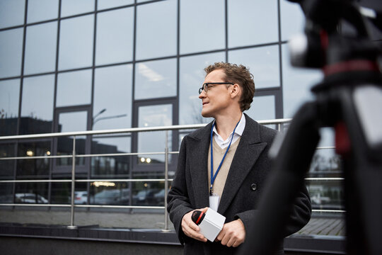 A journalist wearing glasses and a coat engages with a microphone outside an office building, sharing updates.