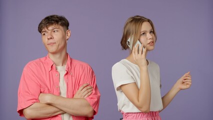 Positive couple of young boyfriend and girlfriend in pink white clothes isolated on purple background, girl talks gossips on smartphone, man waiting gets annoyed.