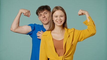 Positive couple of young boyfriend and girlfriend in yellow blue clothes isolated on green background smiling at camera showing strong muscle power arm gesture.