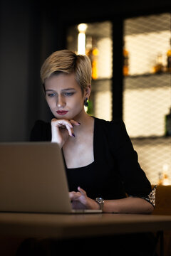 A professional young woman with a modern hairstyle is deeply concentrated while working on her laptop in a dimly lit bar setting.