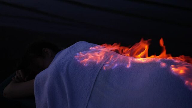 Woman is lying on a massage table with a white towel covering her. Flames appear to be coming from her back as part of a fire therapy treatment