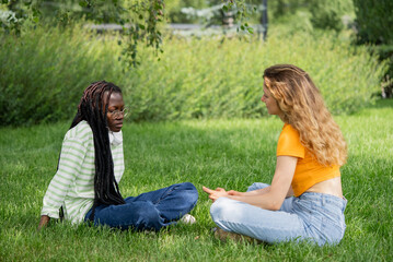 Two young women, possibly students, sitting on grass, having a serious conversation in the park on a sunny day. Engaged, empathetic, and supportive, showing a strong bond of friendship