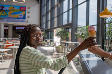 African American woman is reaching for an ice cream cone being handed to her by a food vendor in a brightly lit shopping mall food court