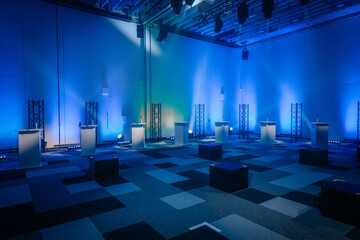 A conference room featuring a row of podiums and microphones, illuminated by blue stage lighting. Geometric carpet pattern adds a contemporary feel. The space is ready for an event or presentation.