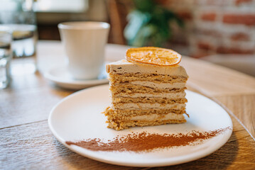 Slice of delicious cake is sitting on a white plate on a wooden table, topped with a slice of dried orange and dusted with cocoa powder