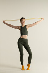 Smiling young woman is exercising with a resistance band. She is dressed in a green sportswear and yellow toe socks. The photo is taken in a minimalistic studio setting with a plain background.