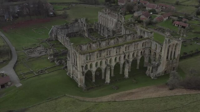 Drone shot of Rievaulx Abbey's ruins surrounded by green landscape and old houses in Yorkshire, England.