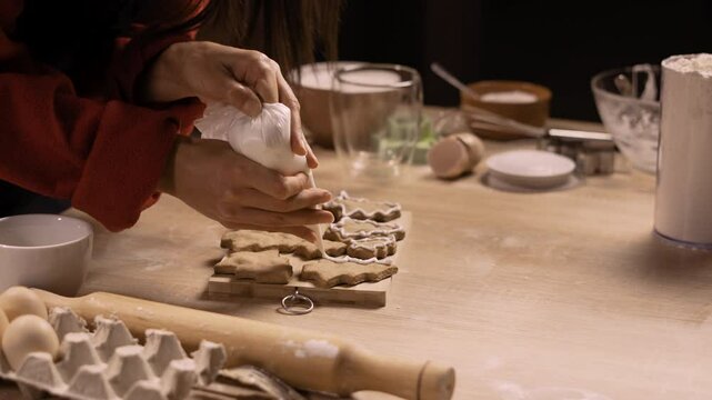 woman decorating gingerbread cookies with white icing using a piping bag, carefully outline shapes on the cookies.