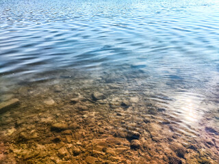 The sunlight reflects on the calm surface of the lake, revealing stones and pebbles underneath the clear water