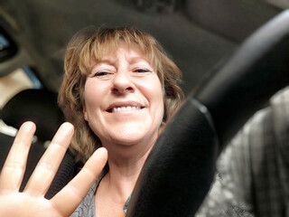 Middle aged woman smiles and waves while sitting in the driver\'s seat of a vehicle during daylight...