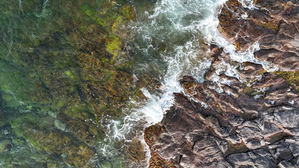 Aerial view of rocky coastline with waves crashing against vibrant seaweed-covered rocks during daylight hours..