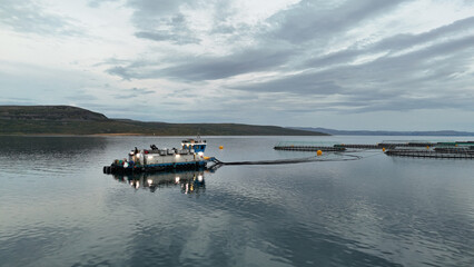 Obraz premium A workboat is involved in aquaculture, floating on calm waters surrounded by mountains and under a cloudy sky.