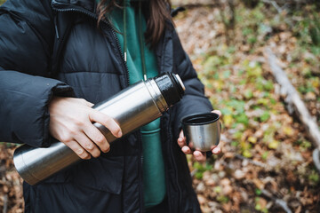 Naklejka premium A joyful woman is seen holding a thermos and a cup, relishing her time outdoors amidst a backdrop of vibrant nature, fully appreciating the stunning beauty that surrounds her