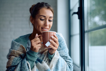 Young woman wrapped in blanket drinking tea by window.