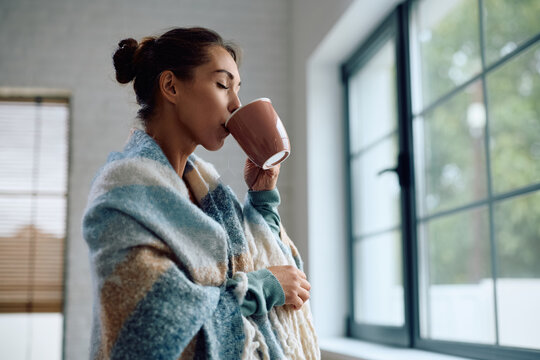 Young woman wrapped in blanket enjoying in cup of warm tea at home.