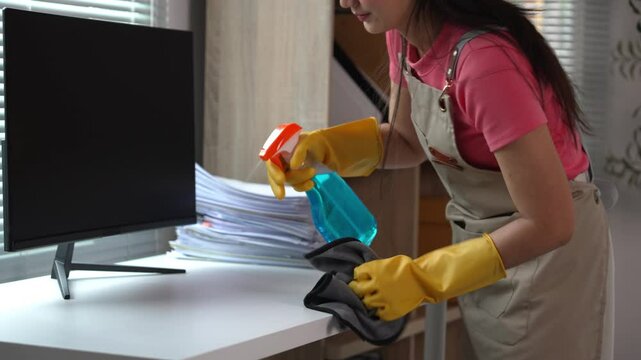 Cleaning lady sanitizes a desk using a spray bottle and microfiber cloth while wearing gloves for effective hygiene