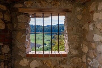 A barred window in a stone wall frames a peaceful view of green hills, fields, and distant forests