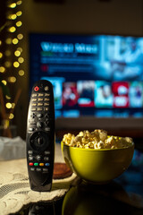 A close-up of a TV remote control on a table with a bowl of popcorn in the foreground, with a TV in the background showing a streaming service interface.