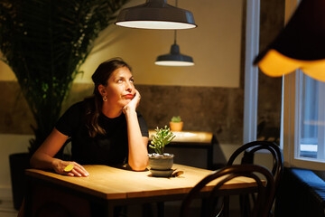 A woman in a black shirt sits at a wooden table, lost in thought, under soft indoor lighting in a tranquil café setting