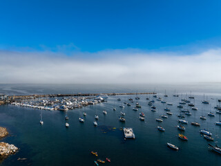 An aerial view captures a marina in Monterey, California, with sailboats, yachts, and kayaks. A long pier extends into the bay, with fog along the coastline.