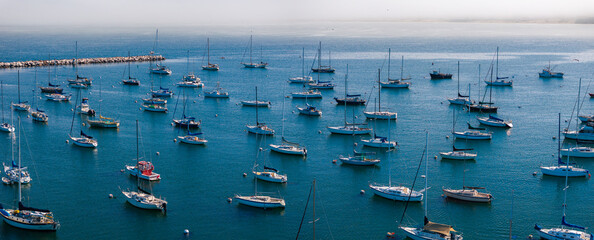 An aerial view captures numerous sailboats and yachts anchored in Monterey's marina, with a breakwater in the background and calm blue waters.